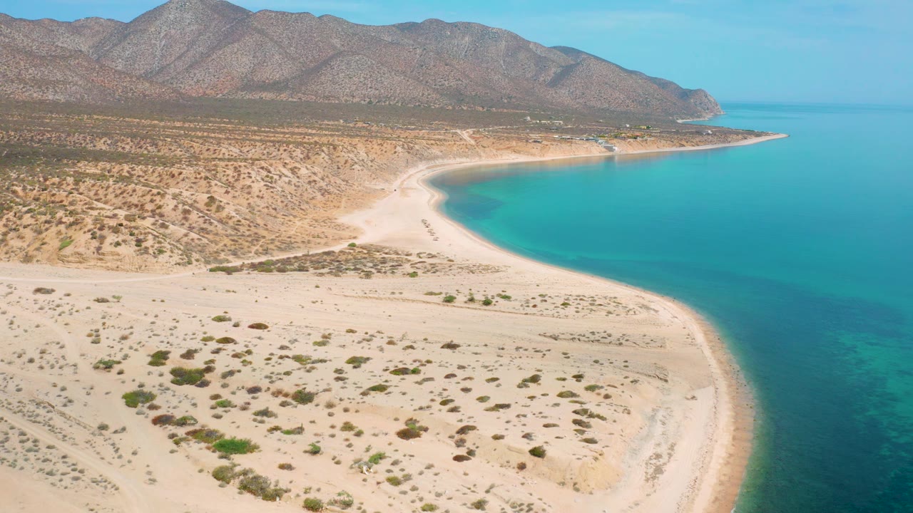 Mid-clip aerial frame over El Sargento La Ventana, Mexico, showing the beach curve from sandy scrub foreground into blue water, with the mountain slope filling the upper background and no visible people.