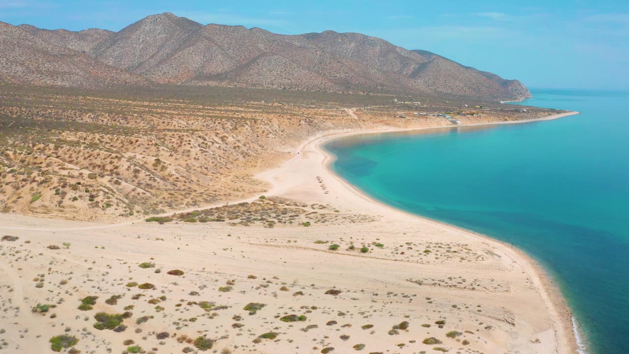 A second aerial view of El Sargento La Ventana shows the sandy foreground, scrub patches, bright blue sea to the right, and a continuous mountain ridge under clear sky, no visible people.