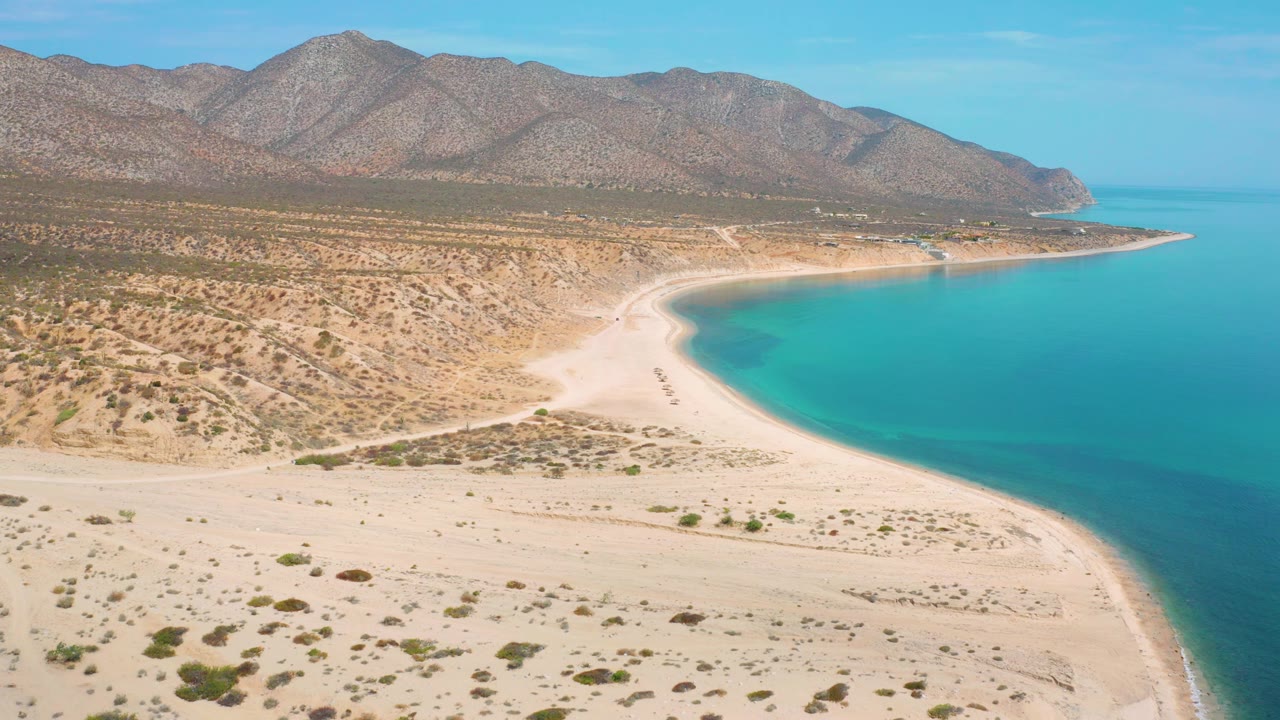 The frame looks across El Sargento La Ventana toward a bright sand spit, sparse desert vegetation, turquoise water along the right side, and rocky mountains beyond, no visible people.