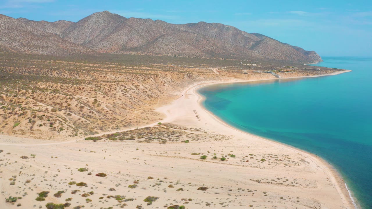 A high aerial frame over El Sargento La Ventana shows broad pale beach in the lower frame, scattered shrubs inland, the bay on the right, and mountains stretching across the top, no visible people.