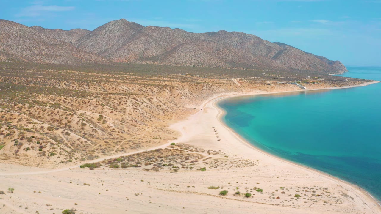 High aerial frame of El Sargento La Ventana, Mexico, showing the turquoise shoreline pushed to the right, open sand below, scrubland across the middle, mountains behind, and no visible people.