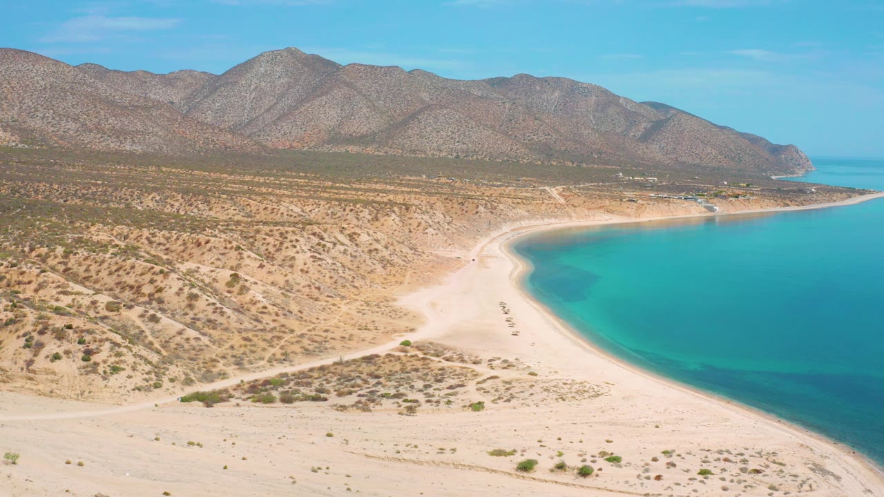 Aerial frame over El Sargento La Ventana, Mexico, with a wider stretch of empty pale sand at the bottom, clear blue-green water at right, desert vegetation inland, rocky hills behind, and no visible people.