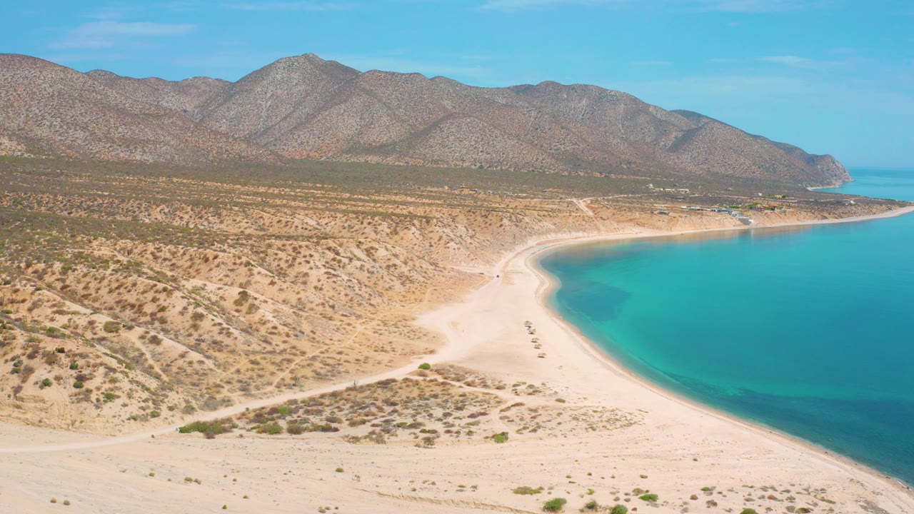 Drone view of a beach curve, bright blue sea, scrub flats, and mountains.