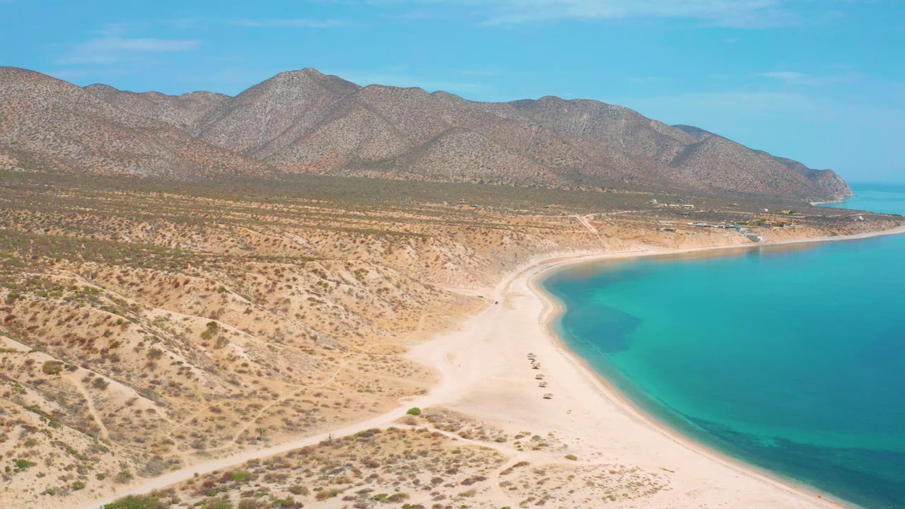 Aerial frame over El Sargento La Ventana, Mexico, with the shoreline slightly farther along the curve, bright sand in the foreground, turquoise sea to the right, mountains beyond, and no visible people.