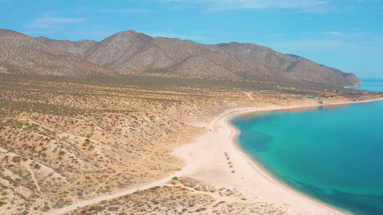 El Sargento La Ventana aerial frame with desert scrub spread across the left side, pale beach and small objects near the waterline, turquoise bay on the right, distant mountains, and no visible people.