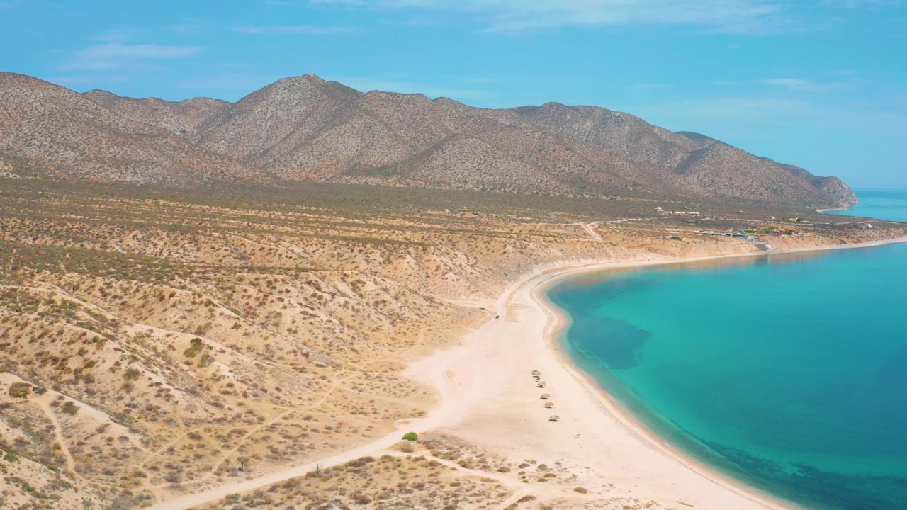 Mid-clip aerial view at El Sargento La Ventana, Mexico, showing more foreground dunes and tracks, a bright sand beach bending around blue ocean, distant mountains, and no visible people.