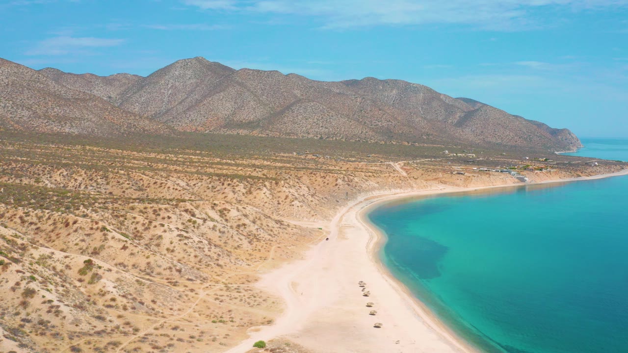 Aerial view of curved white sand beach, turquoise water, desert scrub, and mountains.