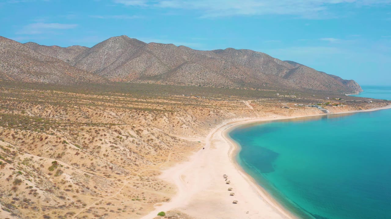 Aerial view in El Sargento La Ventana, Mexico, with the shoreline slightly farther along the frame, turquoise sea on the right, dry brush terrain, mountain backdrop, and no visible people.