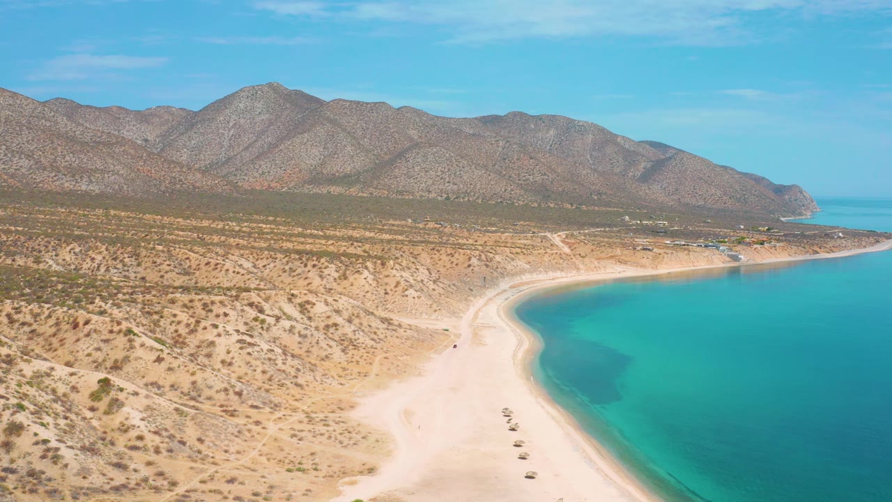 Elevated frame over El Sargento La Ventana, Mexico, showing turquoise water pressing against a pale beach, sparse desert vegetation, rocky mountains, and no visible people.