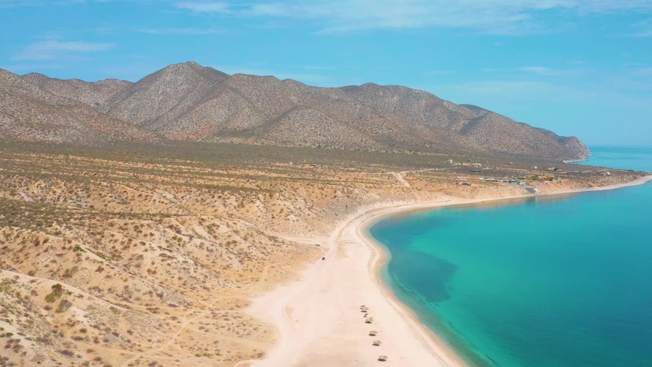 Drone-height view in El Sargento La Ventana, Mexico, with a white sand beach sweeping through the lower frame, turquoise water at right, dry mountains beyond, and no visible people.