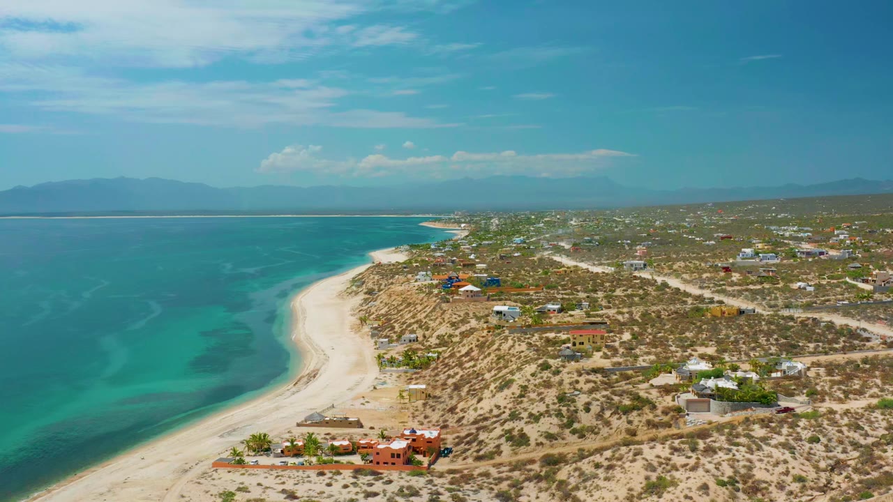 Early aerial frame of El Sargento La Ventana, Mexico, showing shallow blue-green water, a long sand edge, low coastal buildings, scrubland roads, and no visible people.