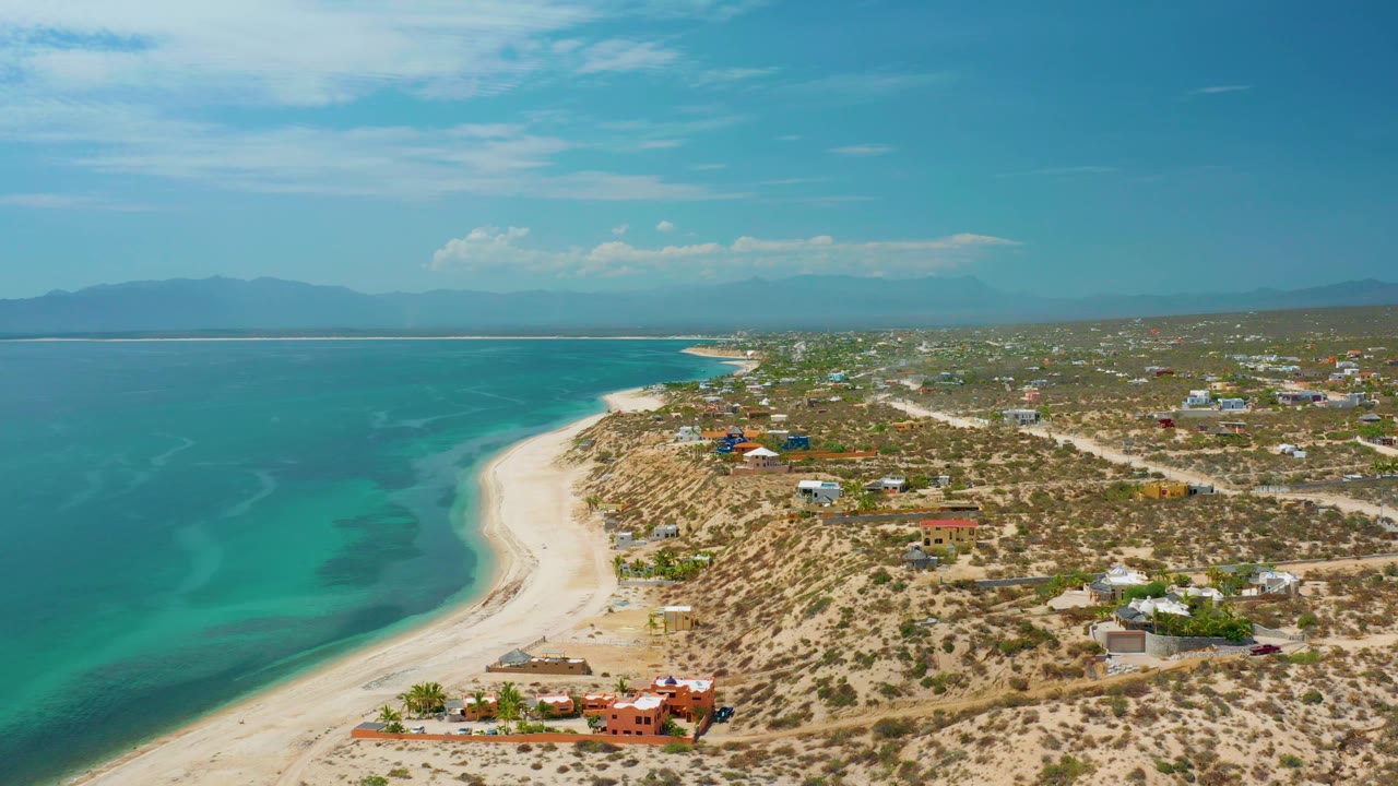 Aerial frame over El Sargento La Ventana, Mexico, with the beach sweeping toward the distance, turquoise sea bands, scattered homes on dry terrain, hazy mountains, and no visible people.
