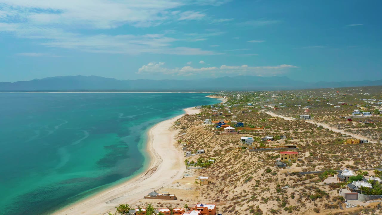 Aerial view over El Sargento La Ventana, Mexico, showing the white shoreline running diagonally past blue-green water, small buildings inland, hazy mountains on the horizon, and no visible people.