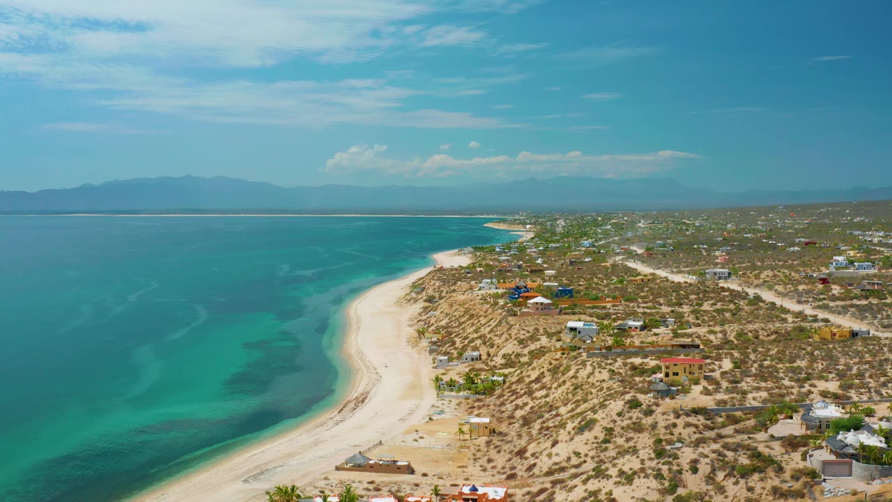 High drone-style frame of El Sargento La Ventana, Mexico, with turquoise bay water, a bright sand beach bending toward the town, scattered rooftops, distant landforms, and no visible people.