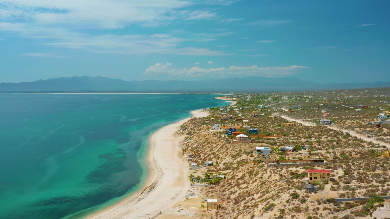 Aerial frame from El Sargento La Ventana, Mexico showing turquoise water on the left, a pale curved beach, dry coastal lots with small buildings, distant mountains, and no visible people.