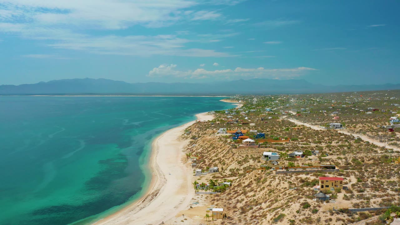 The aerial view over El Sargento La Ventana, Mexico frames shallow turquoise water beside bright sand, with sparse inland buildings, scrub-covered terrain, distant mountains, and no visible people.