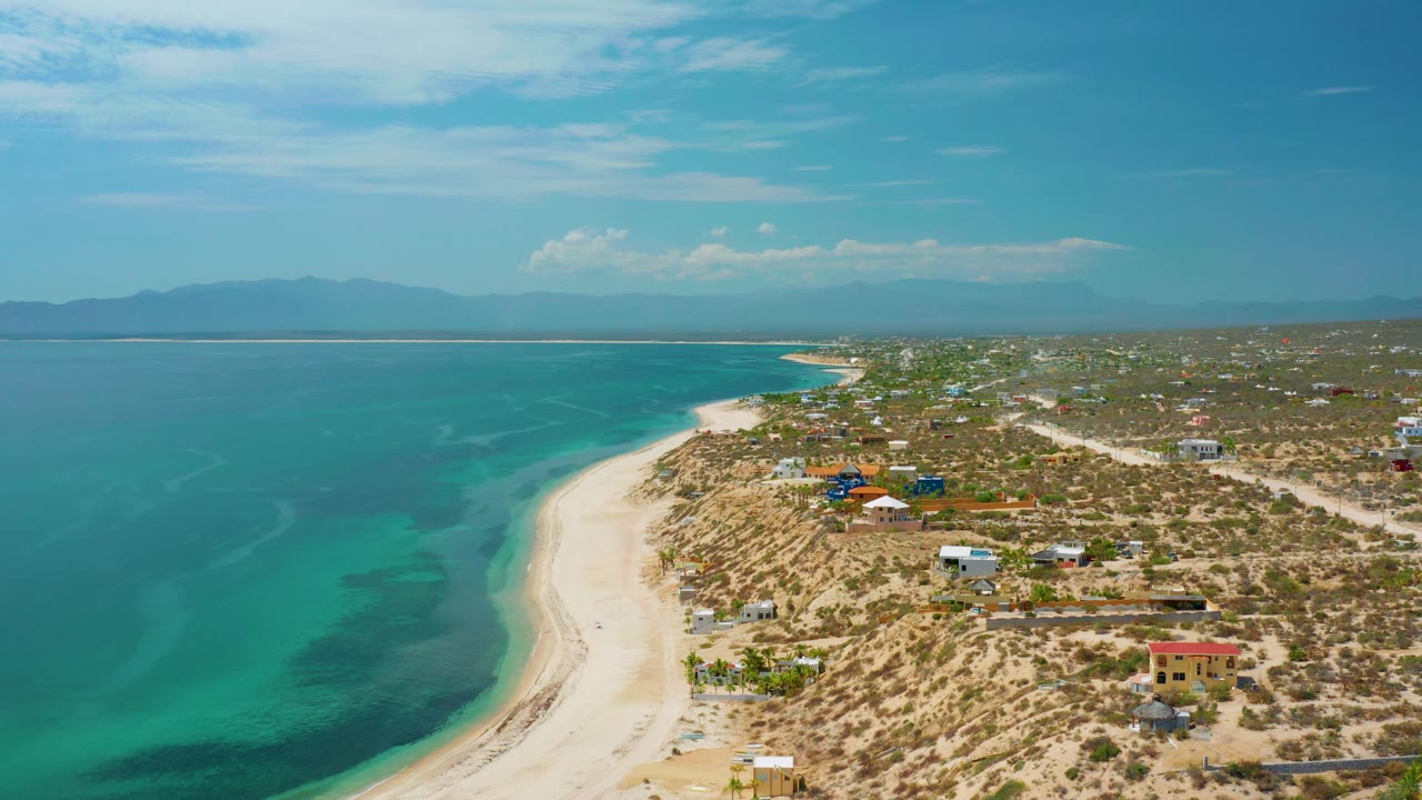 Aerial shoreline with dark sea patches, pale sand curve, low homes, and mountains on the horizon.