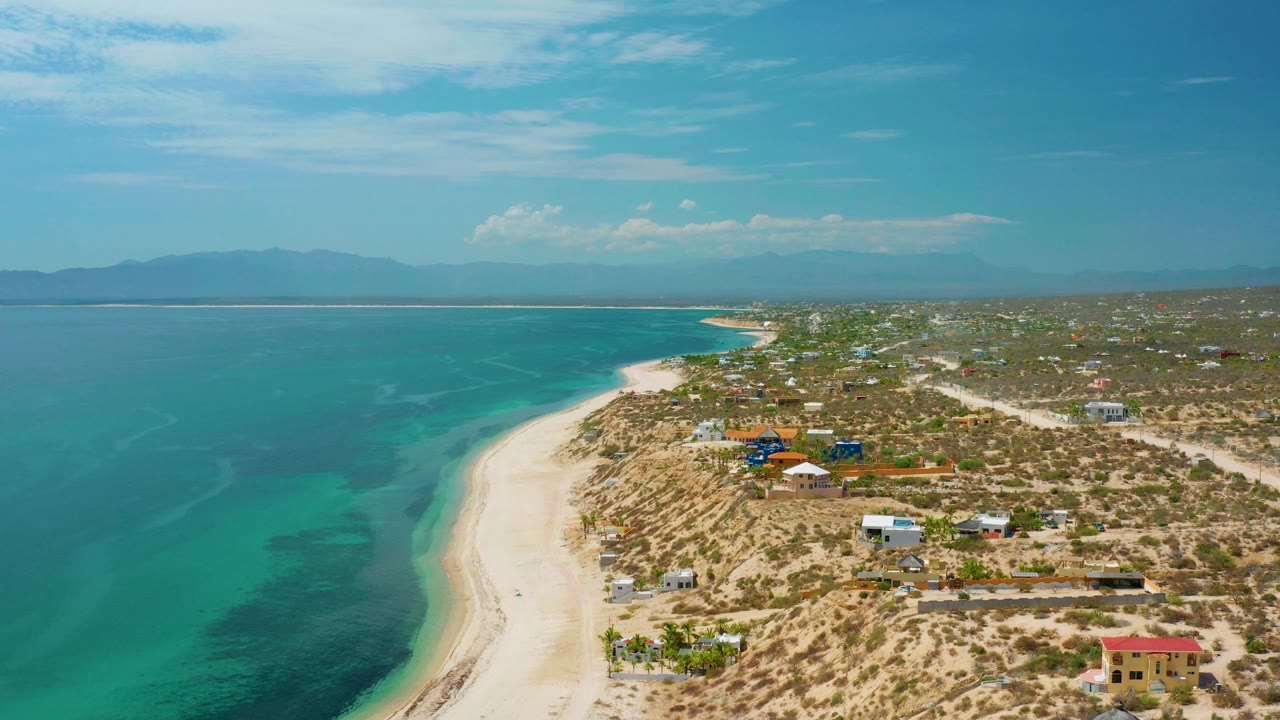Opening aerial view over El Sargento La Ventana, Mexico, with turquoise sea on the left, pale sand beach curving through the frame, scattered buildings on dry land, distant mountains, and no visible people.
