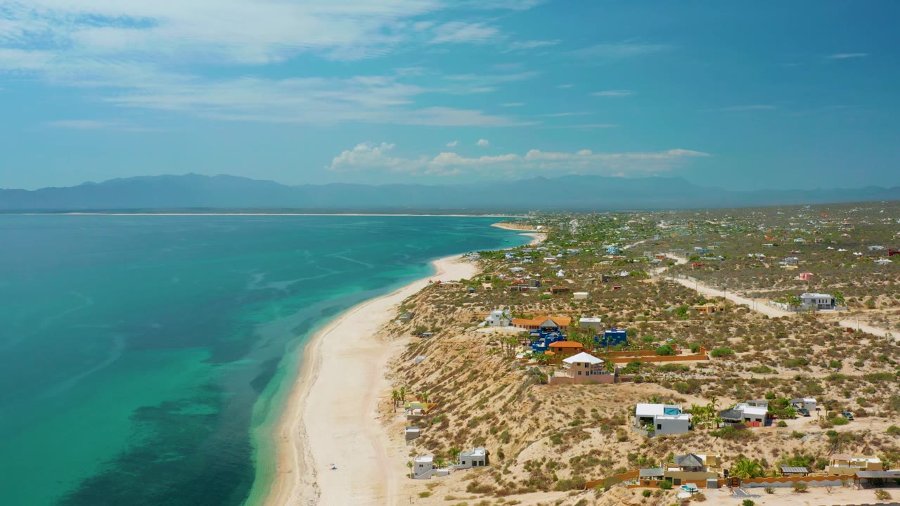 At 0:02, El Sargento La Ventana, Mexico appears from above with the water occupying the left half, the sand line bending inland, dry brush and houses to the right, and no visible people.