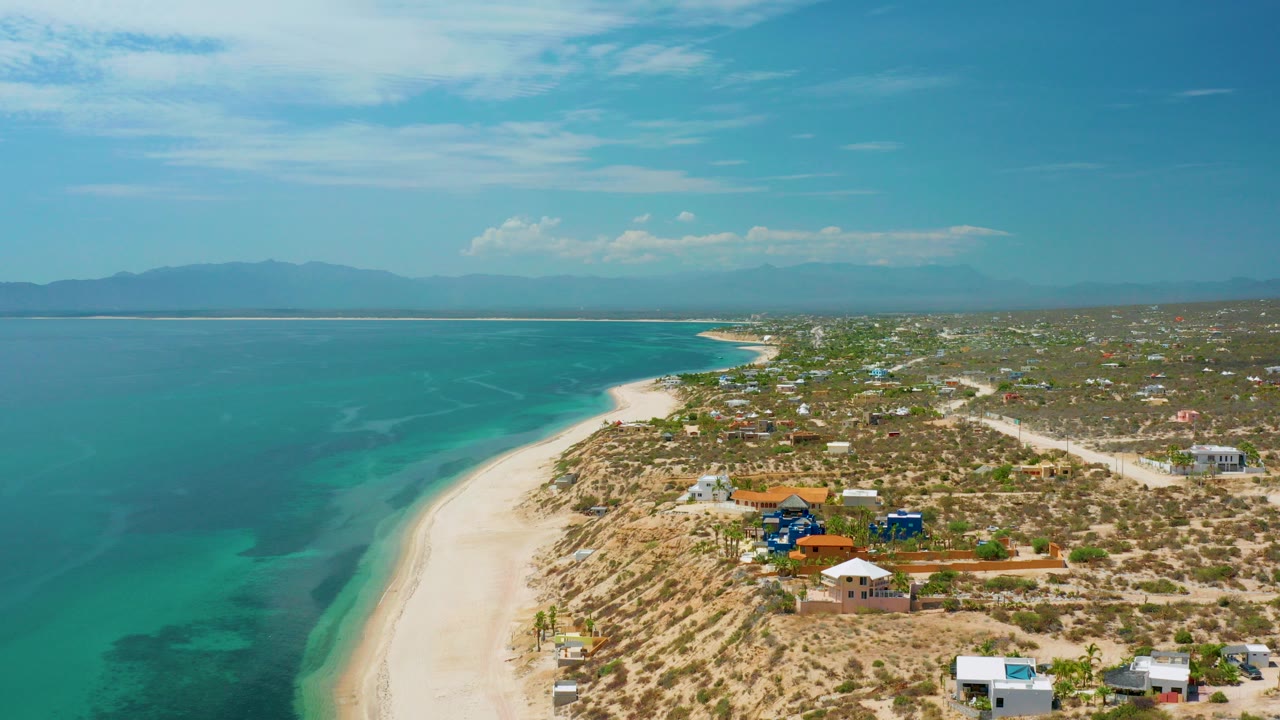 Opening aerial frame over El Sargento La Ventana shows turquoise water beside a pale beach, scattered coastal homes, scrubland, distant mountains, blue sky, and no visible people.