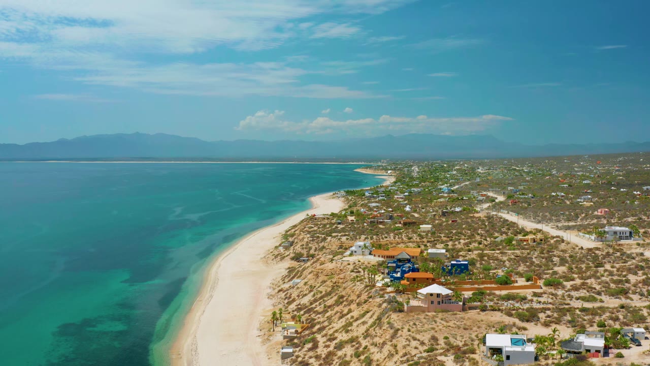 At 0:01 the aerial view holds on El Sargento La Ventana coastline, with more open water filling the left side, a curving sand strip, homes inland, distant mountains, and no visible people.