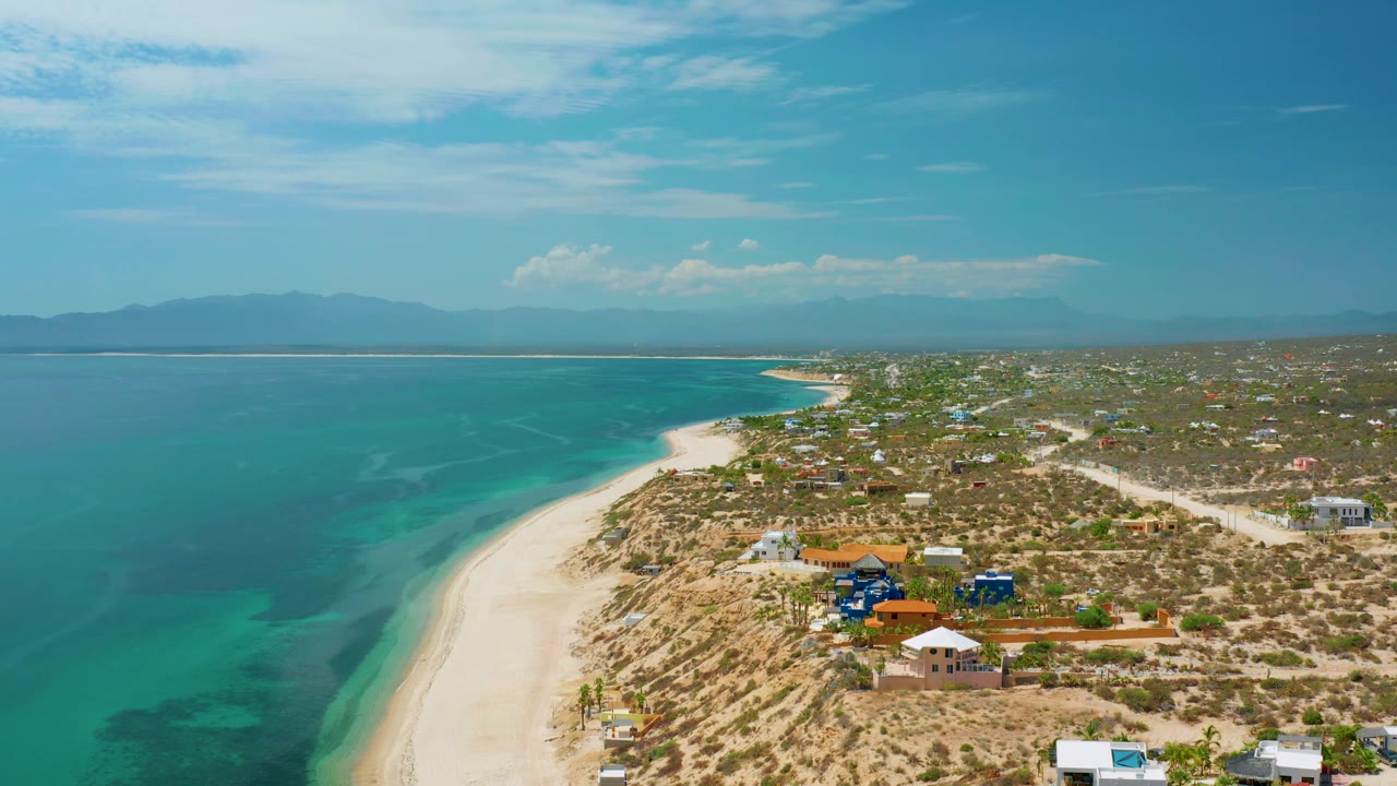 At 0:02 the frame shows turquoise bay water, the sandy beach line, a cluster of colored homes near the shore, scrubby terrain, mountains across the water, and no visible people.