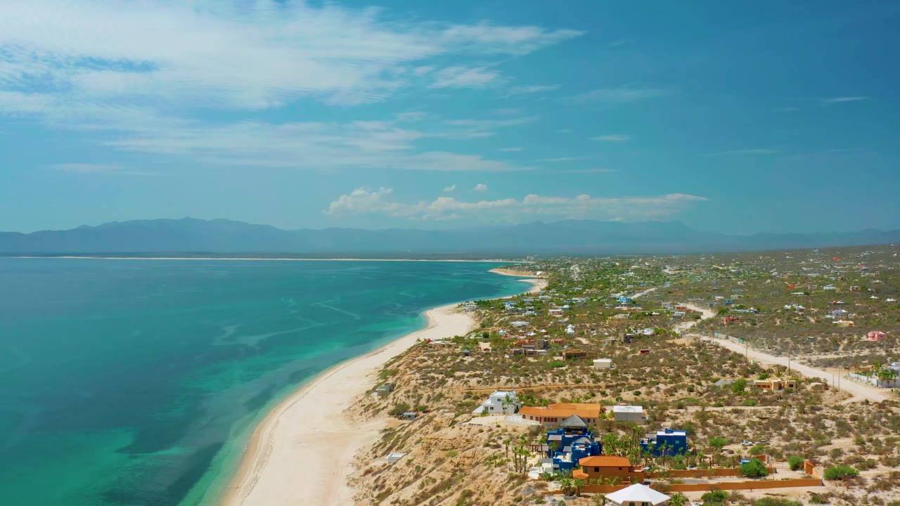 High drone view of El Sargento La Ventana, Mexico, showing blue-green water beside a bright sand curve, sparse coastal homes, dirt roads, hazy mountains, and no visible people.