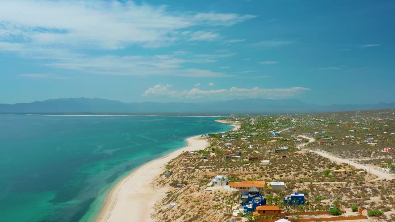 Drone perspective over El Sargento La Ventana, Mexico, with turquoise sea, pale shoreline, a cluster of colorful low buildings near the bottom edge, inland roads, mountains, and no visible people.
