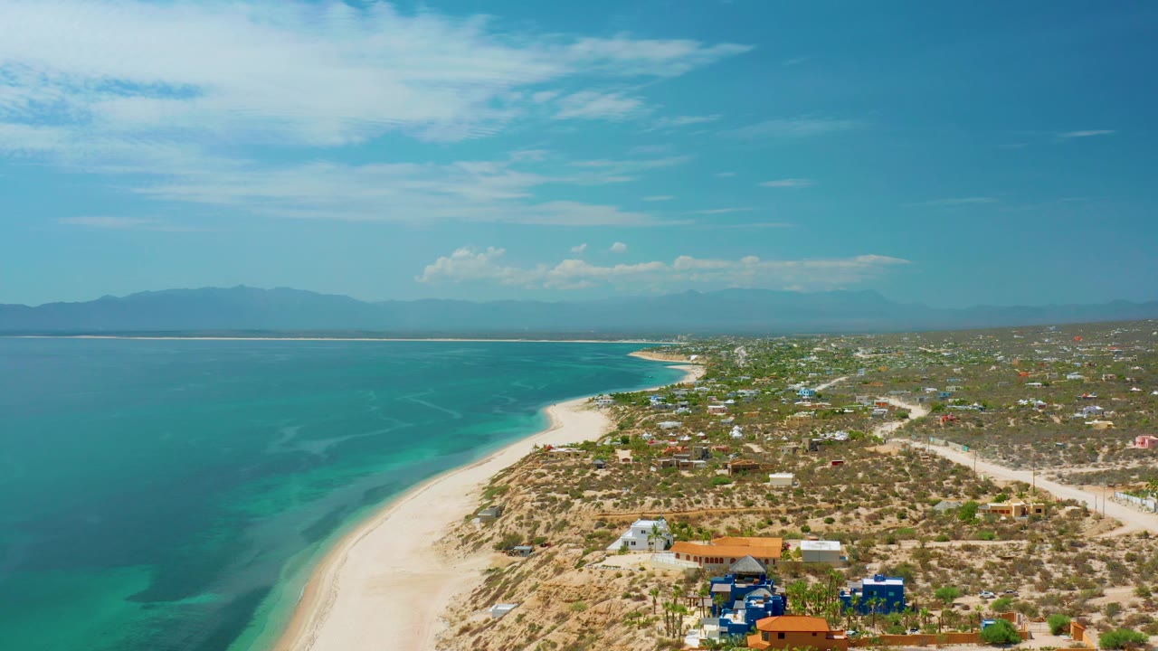 Wide aerial of calm water, white sand, dry coast, and distant mountains.
