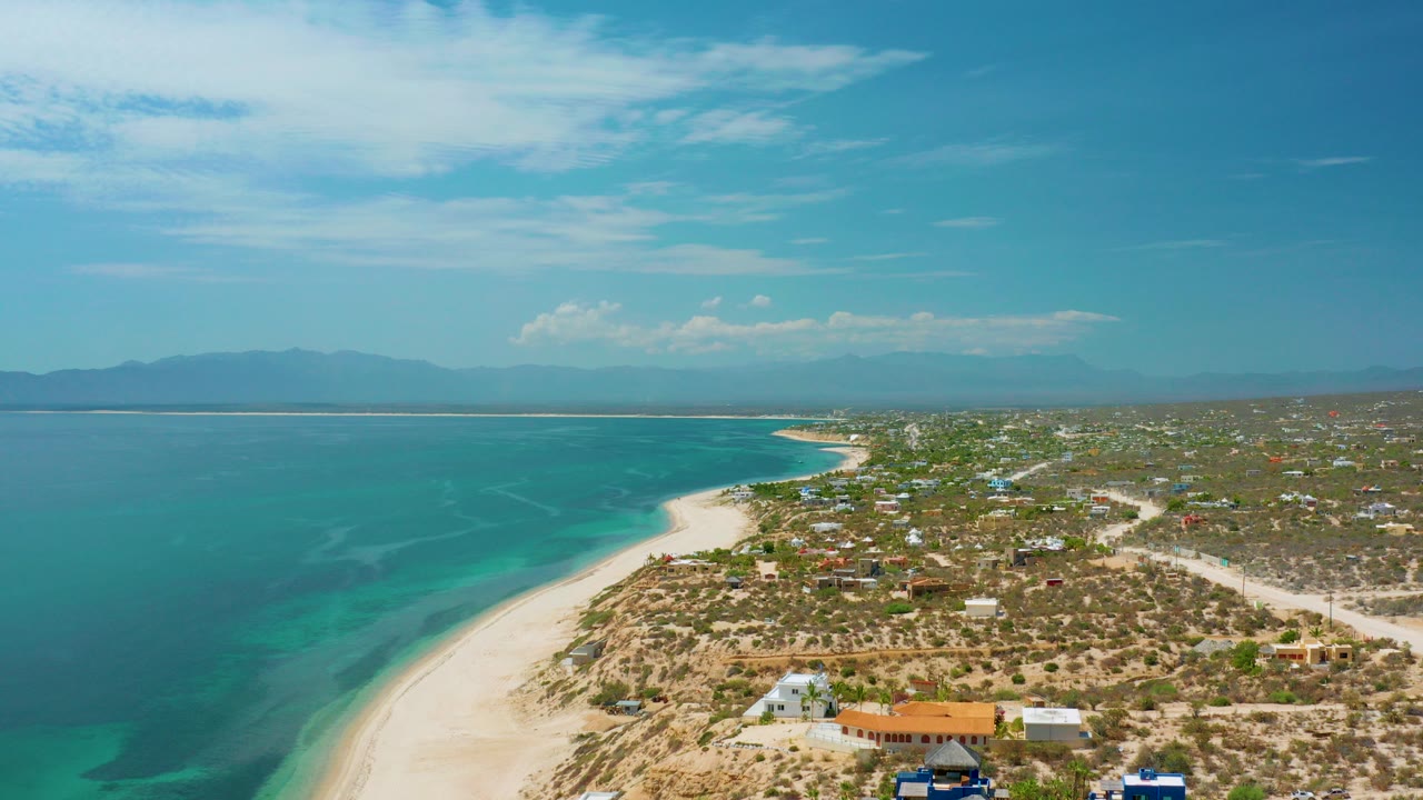 High aerial view over El Sargento La Ventana, Mexico, with turquoise water on the left, a curving sandy beach, scattered low buildings, distant mountains, and no visible people.