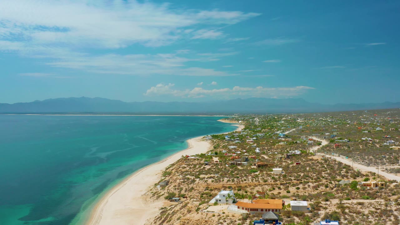 Aerial view of El Sargento La Ventana, Mexico, with the shoreline running diagonally through frame, turquoise sea to the left, low coastal buildings to the right, and no visible people.