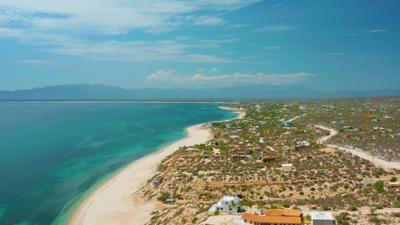 Opening aerial view over El Sargento La Ventana, Mexico, with turquoise sea at left, pale beach curving through the center, scattered buildings on dry land, distant mountains, and no visible people.