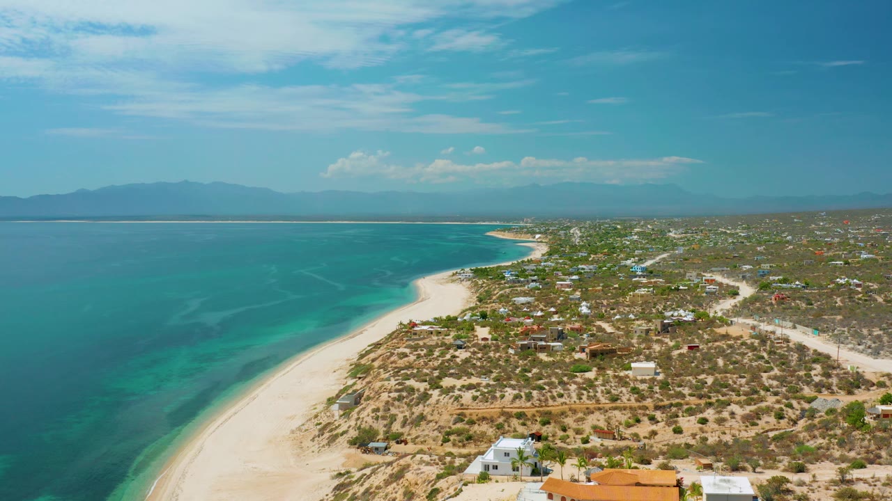 Drone-height view over El Sargento La Ventana, Mexico, with turquoise water spreading across the left side, a sandy beach edge, sparse homes and roads to the right, hazy mountains, and no visible people.