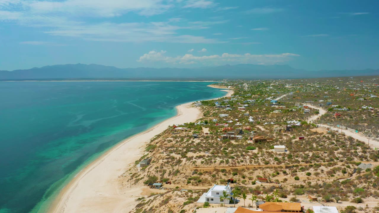 Second aerial frame of El Sargento La Ventana, Mexico, showing the sea occupying the left side, a long sand edge, scattered homes and roads inland, hazy mountains beyond, and no visible people.