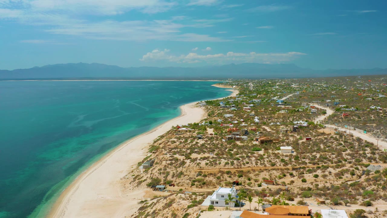 Third aerial frame over El Sargento La Ventana, Mexico, with clear water patterns near shore, a broad sandy strip, clustered low buildings inland, distant mountain silhouettes, and no visible people.