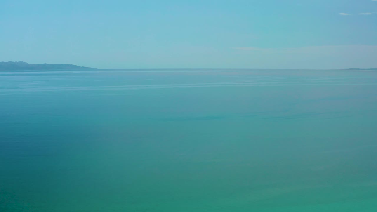 Blue green ocean water stretches toward low land on the horizon in El Sargento La Ventana, Mexico, with faint darker patches under the surface and no visible people.