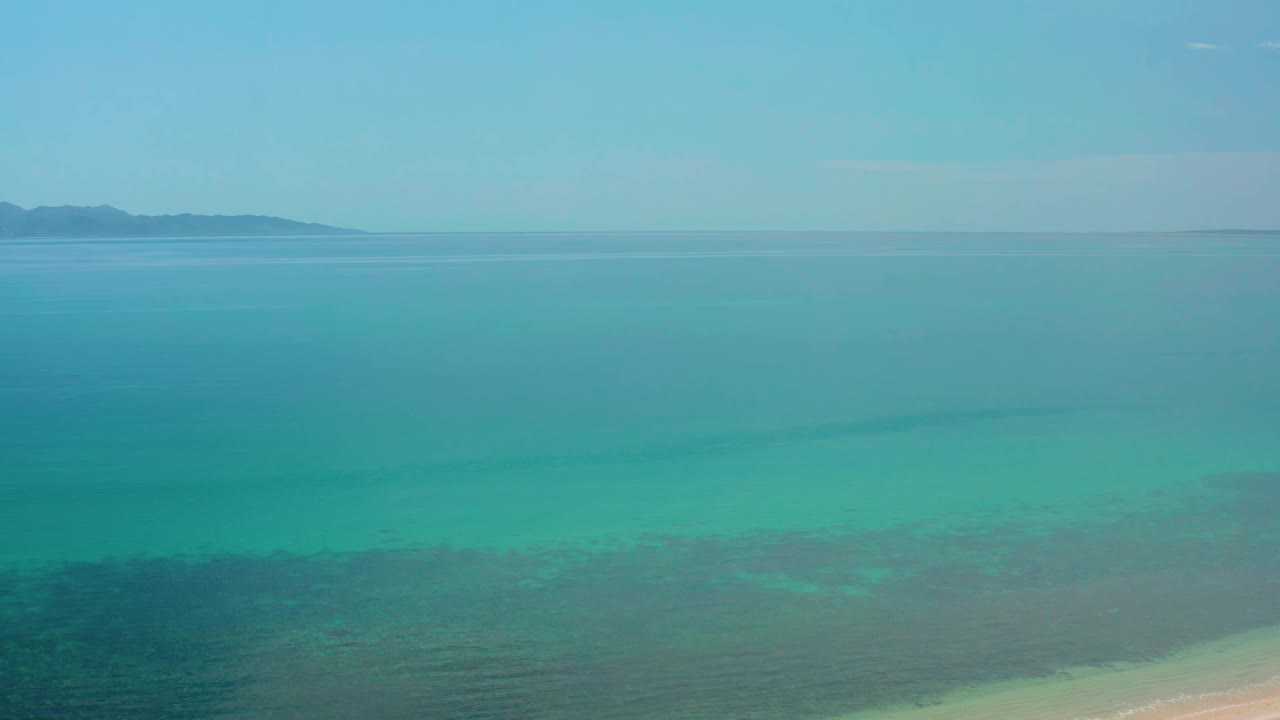 Aerial frame over El Sargento La Ventana, Mexico, with turquoise sea filling most of the view, a thin strip of pale sand at lower right, distant land at upper left, and no visible people.