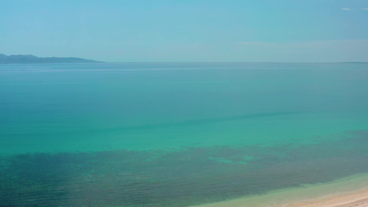 Aerial frame over El Sargento La Ventana, Mexico, showing a broad field of blue-green water, faint darker bands beneath the surface, low land across the horizon, and no visible people.