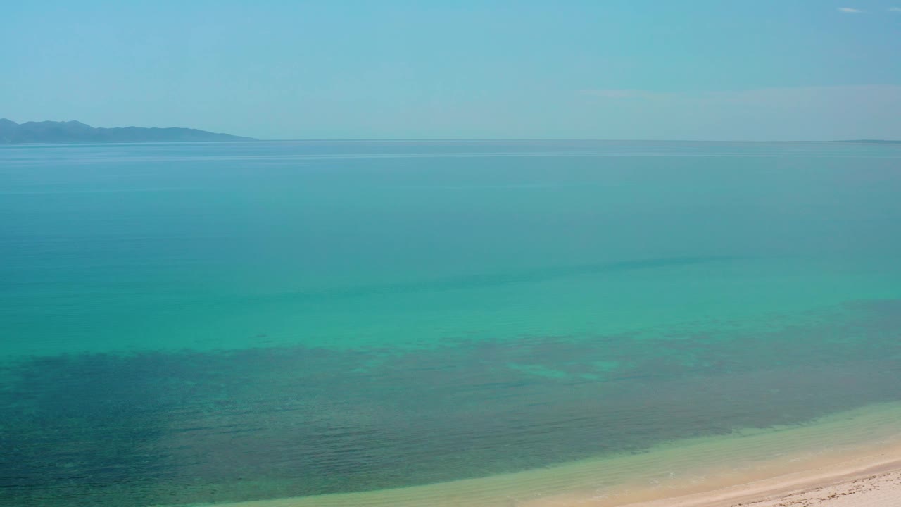 Mid-clip aerial frame over El Sargento La Ventana, Mexico, with open turquoise water, darker reef-like or seaweed-like patches, distant land at the left horizon, and no visible people.