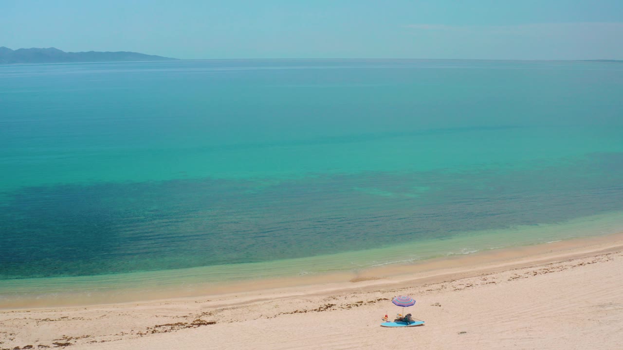 A high aerial frame over El Sargento La Ventana, Mexico shows pale sand, turquoise shallows, darker blue water bands, distant land on the horizon, and a small blue-purple umbrella setup near the lower center.