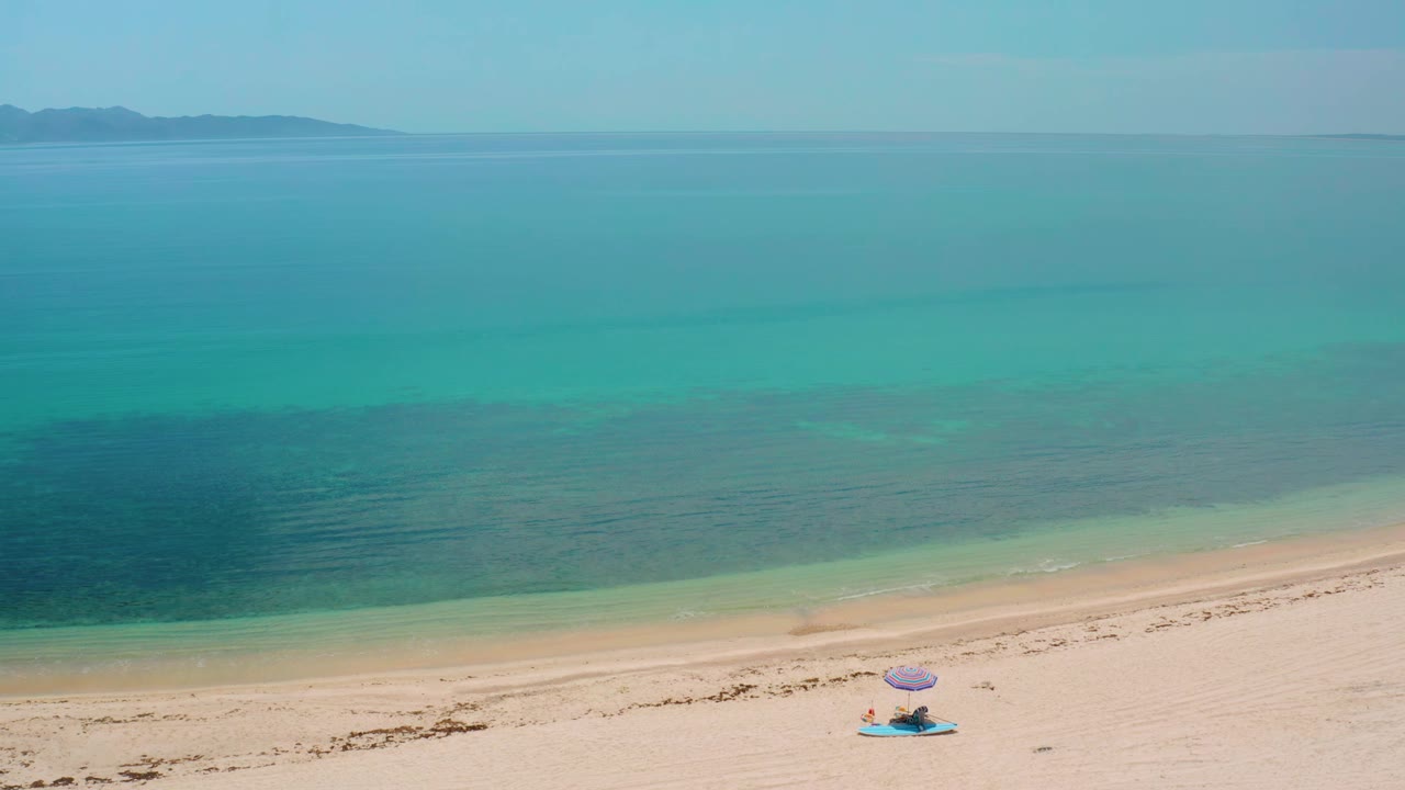 High view of a quiet shoreline with a beach umbrella and towel below clear blue water.