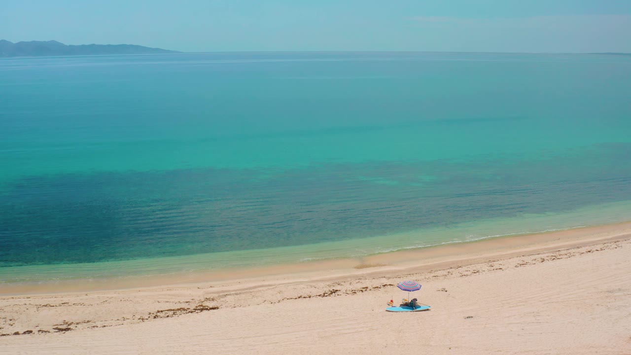 A wide aerial view in El Sargento La Ventana, Mexico places the small umbrella setup toward the lower right, with open sand, turquoise shallows, deeper water, and distant land across the horizon.
