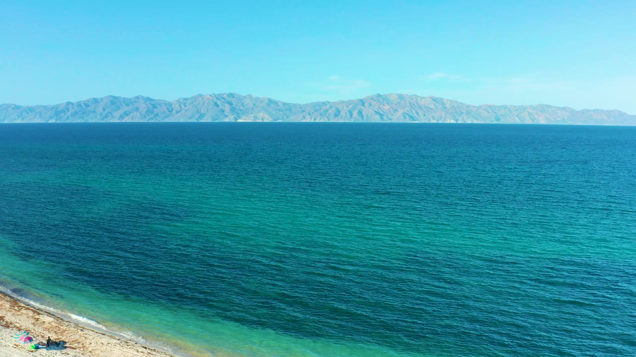 A slightly lower view includes more rocky shoreline at the lower left, teal water shifting to deeper blue, mountains across the distance, and no visible people in El Sargento La Ventana, Mexico.