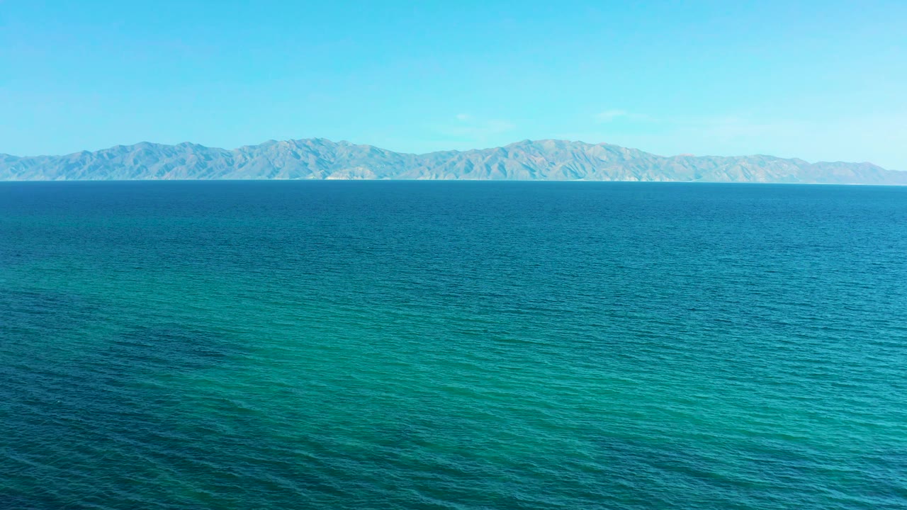 Aerial view over blue-green water at El Sargento La Ventana, Mexico with a pale mountain range in the distance and no visible people or structures.