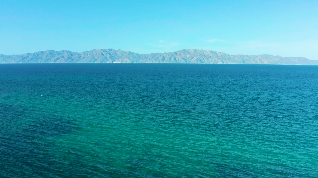 Wide aerial frame of El Sargento La Ventana, Mexico showing layered blue ocean tones, a continuous distant mountain ridge, clear daylight, and no visible people.