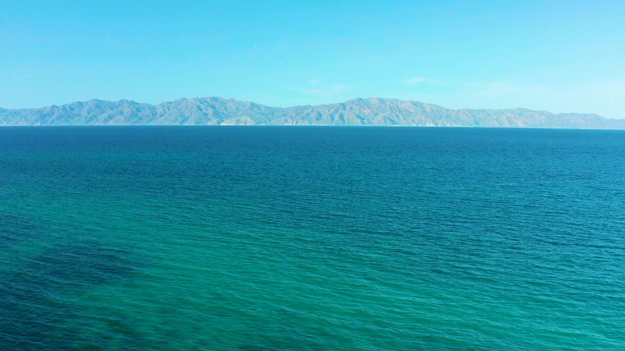 Mid-clip aerial view of calm turquoise sea in El Sargento La Ventana, Mexico, with low mountains across the horizon and no visible people, boats, or buildings.