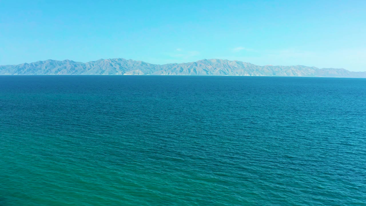 At 0:01, turquoise water texture fills the lower frame while the El Sargento La Ventana mountain horizon remains level beneath a bright sky, with no visible people.