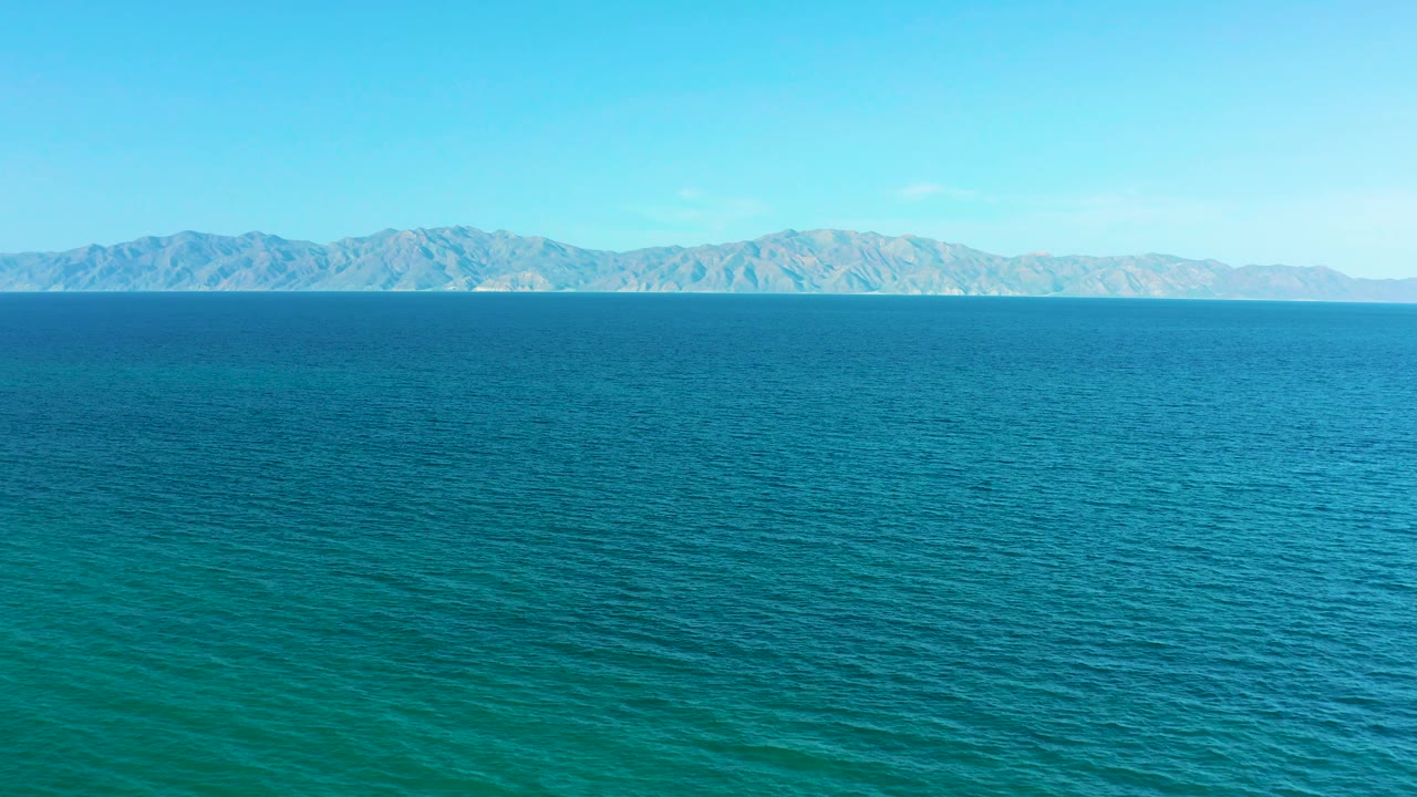 At 0:02, the frame holds a wide band of open blue sea and a continuous distant mountain range near El Sargento La Ventana, Mexico, with no visible people.