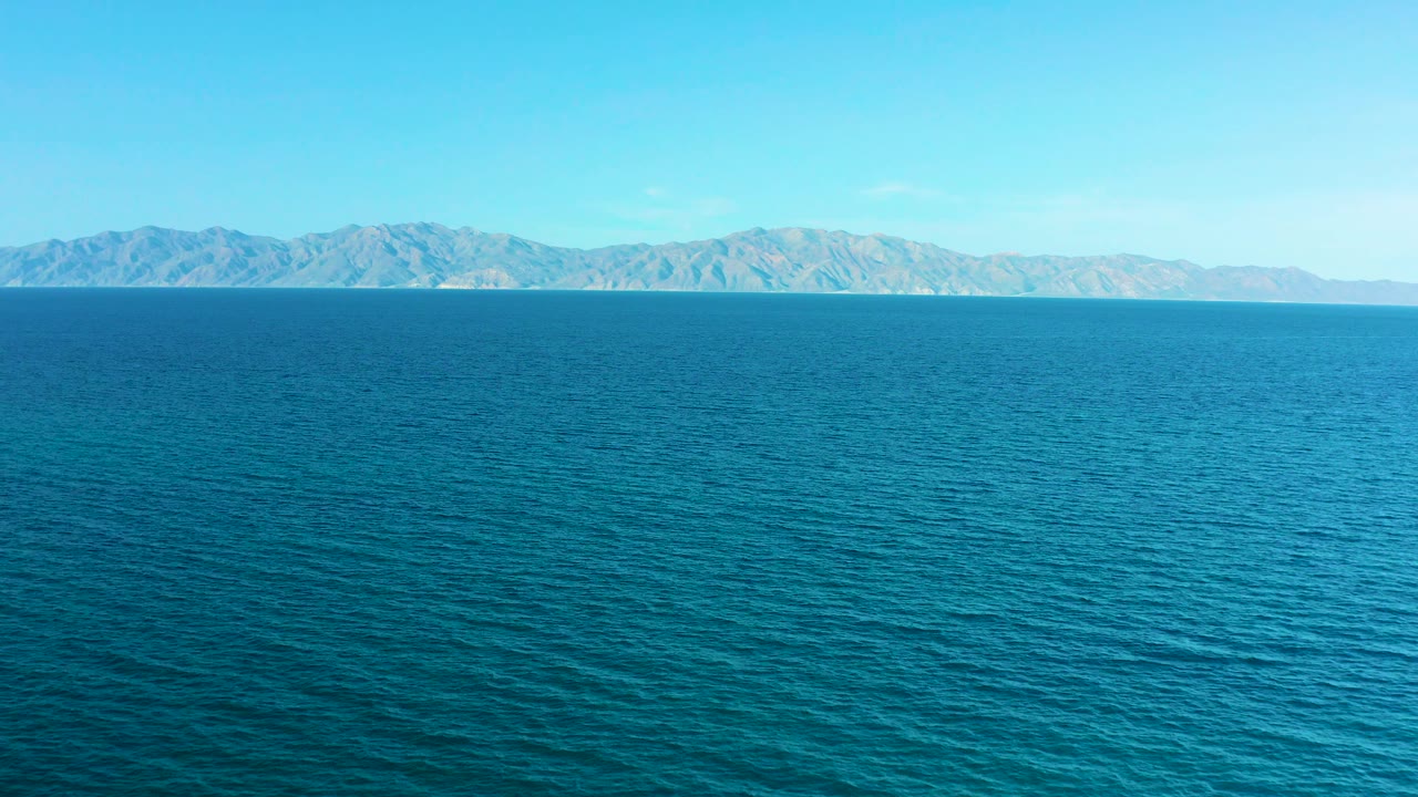 Blue sea occupies the foreground in El Sargento La Ventana, Mexico, with pale mountains running across the distant coastline and no visible people.