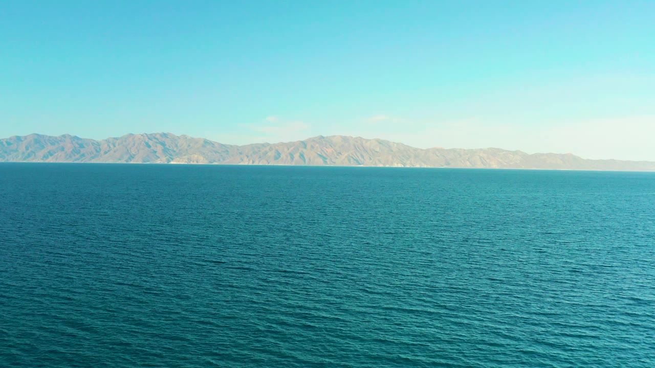 Opening frame of blue ocean in El Sargento La Ventana, Mexico, with rippled water in front, tan mountains on the horizon, clear sky above, and no visible people.